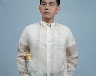 May include: Off-white long-sleeved shirt with intricate embroidery details. The shirt features a mandarin collar and button-down front. The brand logo "Aquino's Artisan Embroidery" is visible on the left side. The model is wearing black pants.