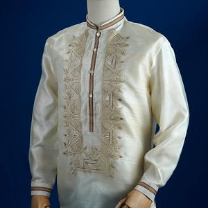 以下が含まれることがあります： 前面と袖口に複雑な刺繍が施された、オフホワイトの長袖シャツ。 シャツはマンダリンカラーとボタンダウンフロントを備えています。 刺繍は対照的な茶色です。 シャツはマネキンに展示されています。