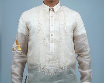 May include: Off-white formal shirt with intricate embroidery down the front and on the sleeves. The shirt has a mandarin collar and button-down closure. The sleeves are semi-sheer. The shirt is worn with black pants. The logo "Aquino's Artisan Embroidery" is visible.