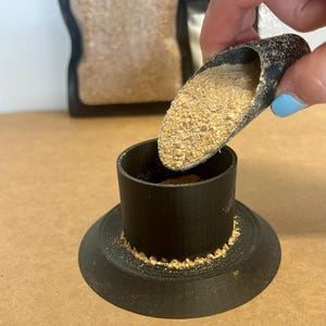 Puede incluir: Un embudo negro impreso en 3D con una base ancha y una parte superior cilíndrica, que se llena con una sustancia granular de color marrón claro de una pequeña cuchara. Una bolsa de la misma sustancia está en el fondo.