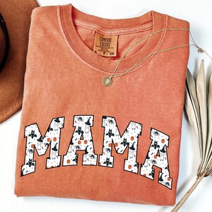 May include: An orange Comfort Colors t-shirt with the word "MAMA" in a white font filled with Halloween-themed patterns. A gold necklace with a pendant is draped across the shirt. A brown hat is visible in the upper left corner.