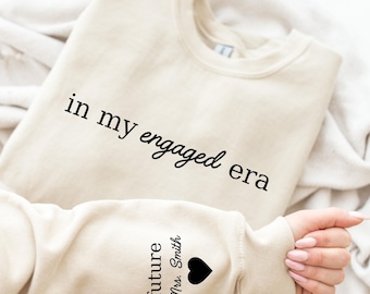 May include: A beige sweatshirt with the text "in my engaged era" printed in black. The sleeve features the words "future Mrs. Smith" and a black heart. The sweatshirt is folded on a white surface.