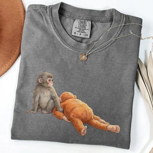 May include: A gray Comfort Colors t-shirt featuring a watercolor illustration of two monkeys. One monkey is sitting, and the other is lying down. A silver necklace with a small pendant is draped on the shirt.