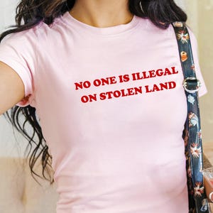 May include: A light pink t-shirt with the text "NO ONE IS ILLEGAL ON STOLEN LAND" in red. The shirt is short-sleeved and form-fitting. The model is wearing blue jeans and carrying a black purse with a floral pattern.