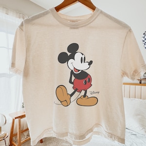 May include: Cream-colored t-shirt featuring a classic Mickey Mouse graphic. The cartoon character is depicted in black, red, and tan, with the Disney logo printed below. The shirt is hanging on a wooden hanger.