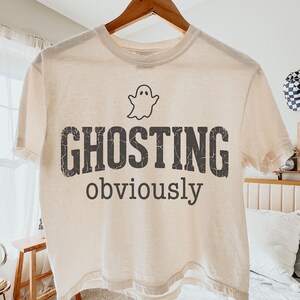 May include: A cream-colored short-sleeve t-shirt featuring a black graphic of a small ghost icon above the distressed, bold, capitalized text GHOSTING and the lowercase word obviously printed in a simple, dark gray sans-serif font.