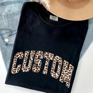 May include: Black t-shirt with the word "CUSTOM" in a leopard print design. The shirt is folded on top of a pair of light blue jeans and a tan hat. A gold necklace is visible.