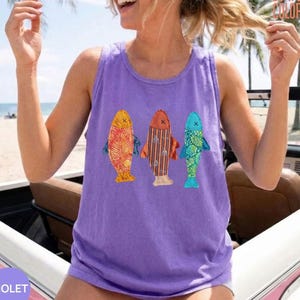 May include: A light purple sleeveless tank top featuring a graphic print of three stylized fish. The left fish is orange with a leaf pattern, the center fish is brown with vertical stripes, and the right fish is teal with a geometric pattern.