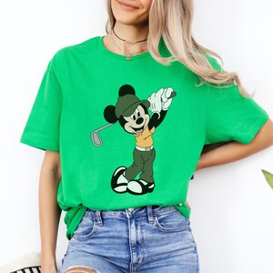 May include: Bright green t-shirt featuring a cartoon Mickey Mouse character playing golf. Mickey is wearing a green hat, khaki shirt, and green pants. The golf club is silver. The shirt is a solid, vibrant green color.