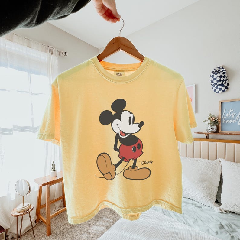 May include: Yellow short-sleeved t-shirt featuring a graphic of Mickey Mouse. The cartoon character is depicted in black, red, white, and tan colors. The Disney logo is printed in black on the lower right side of the shirt.
