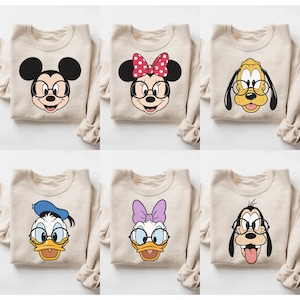 May include: A collection of six beige crewneck sweatshirts, each featuring a different Disney character graphic. The characters include Mickey Mouse, Minnie Mouse, Pluto, Donald Duck, Daisy Duck, and Goofy, all wearing round black glasses.