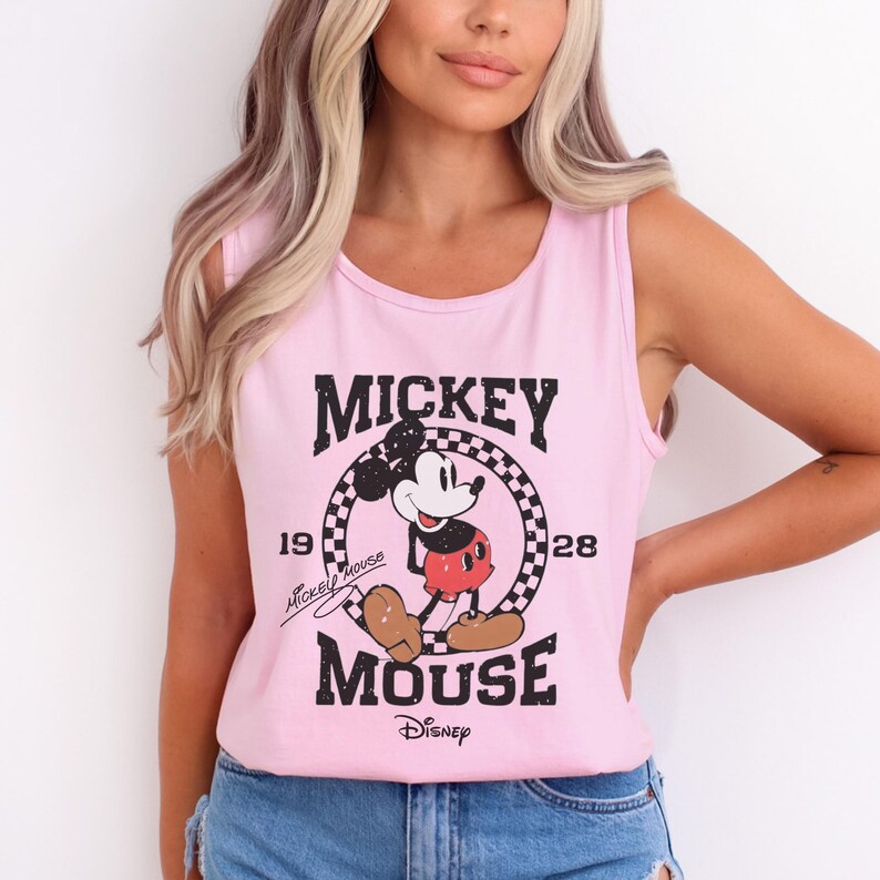 Pu&ograve; includere: Canotta rosa chiaro con grafica di Topolino. Il design include la parola "Mickey" sopra un motivo a scacchiera circolare con Topolino al centro. Le parole "Mouse" e "Disney" sono in basso.