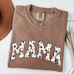 May include: A brown Comfort Colors t-shirt with the word "MAMA" in white block letters. The letters are filled with a pattern of Halloween-themed images, including bats, spiders, and pumpkins. A silver necklace with a pendant is draped across the shirt.