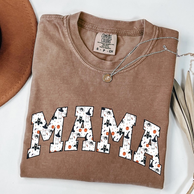May include: A brown Comfort Colors t-shirt with the word "MAMA" in white block letters. The letters are filled with a pattern of Halloween-themed images, including bats, spiders, and pumpkins. A silver necklace with a pendant is draped across the shirt.