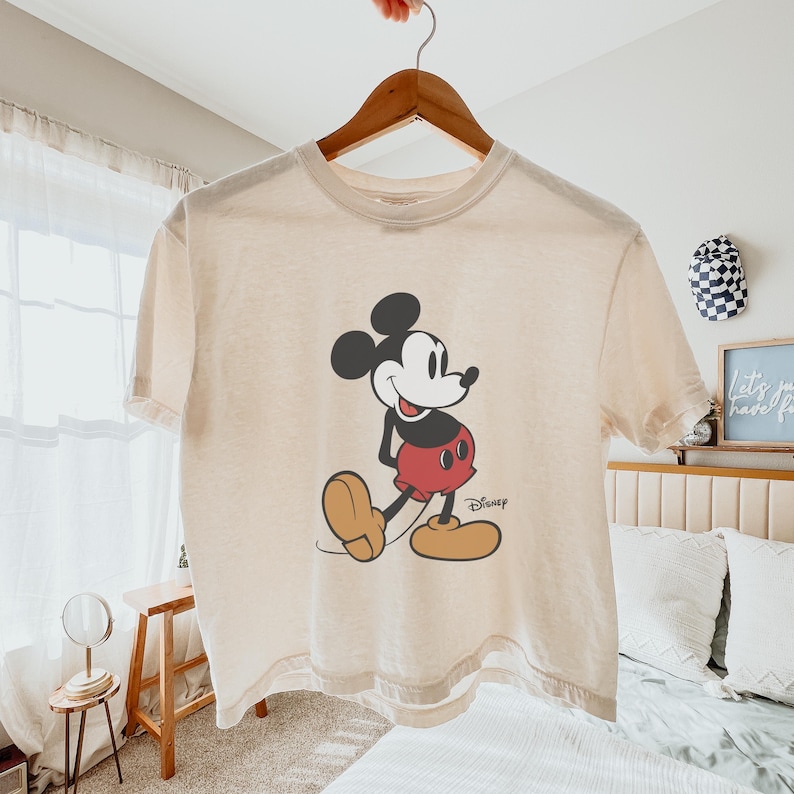 May include: Cream-colored t-shirt featuring a classic Mickey Mouse graphic. The cartoon character is depicted in black, red, and tan, with the Disney logo printed below. The shirt is hanging on a wooden hanger.