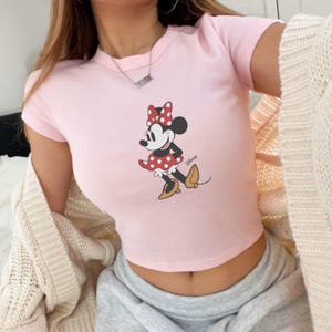 Pode incluir: Top cropped rosa claro com uma estampa da Minnie Mouse. Minnie est&aacute; vestida com um vestido de bolinhas vermelhas, la&ccedil;o combinando e sapatos amarelos. O logotipo da Disney &eacute; vis&iacute;vel no canto inferior direito.