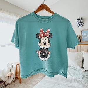 Pode incluir: Camiseta azul-petr&oacute;leo com um desenho da Minnie Mouse. O desenho mostra a Minnie Mouse com um la&ccedil;o vermelho de bolinhas brancas, orelhas pretas e uma roupa preta. A camisa est&aacute; pendurada em um cabide de madeira.