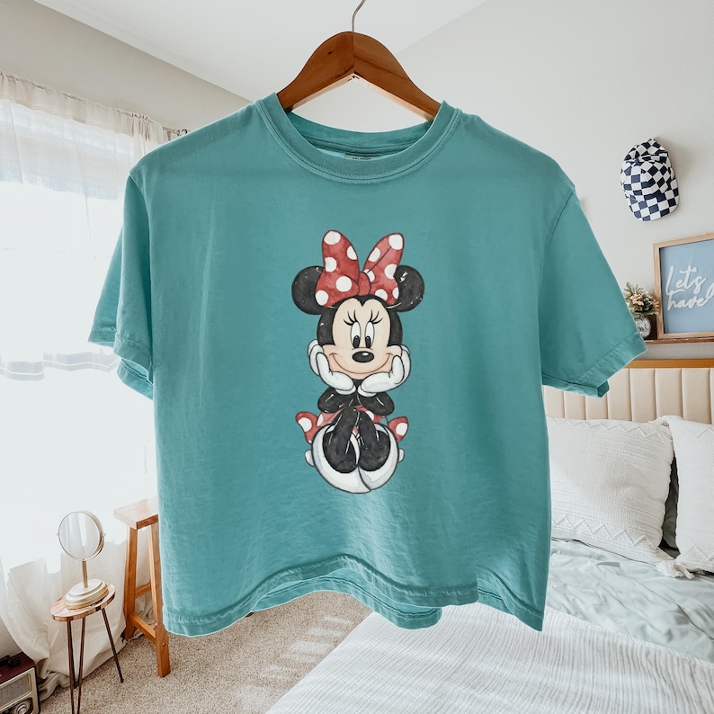 Pode incluir: Camiseta azul-petr&oacute;leo com um desenho da Minnie Mouse. O desenho mostra a Minnie Mouse com um la&ccedil;o vermelho de bolinhas brancas, orelhas pretas e uma roupa preta. A camisa est&aacute; pendurada em um cabide de madeira.