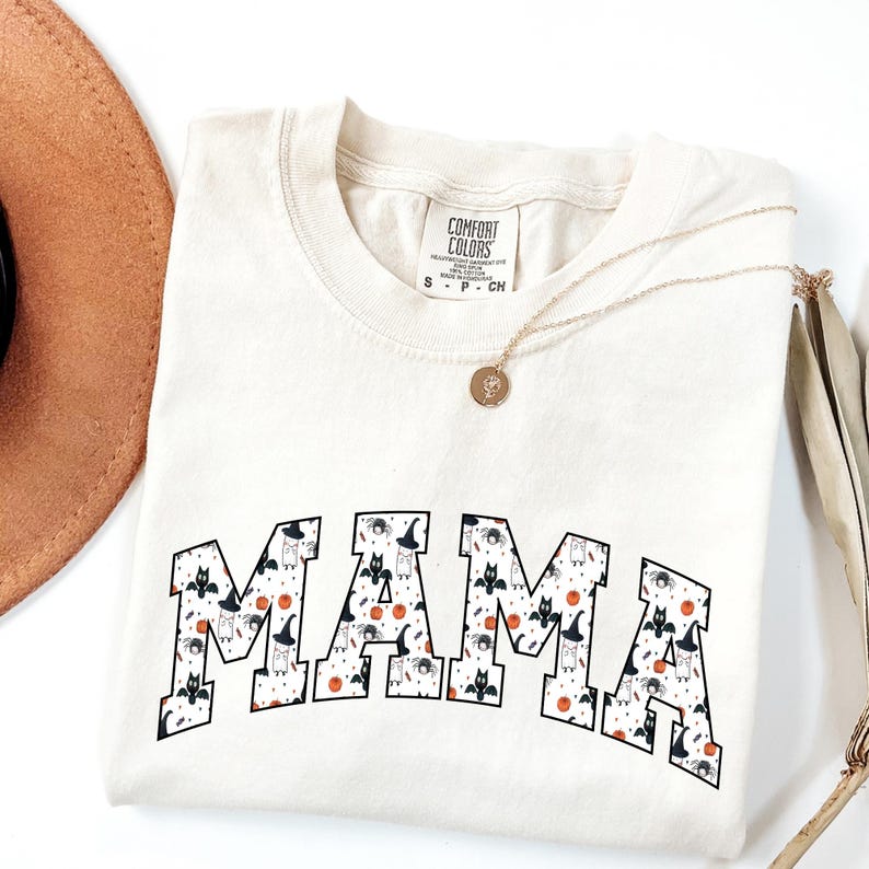 May include: Cream-colored t-shirt with the word "MAMA" in a bold, patterned font featuring Halloween-themed illustrations. The shirt is folded, with a gold necklace and a brown hat visible. The shirt is made by Comfort Colors.