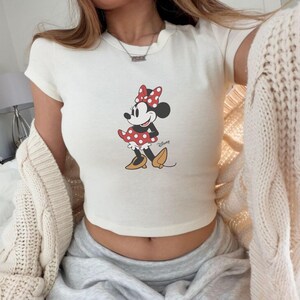 Pode incluir: Top cropped creme com um gr&aacute;fico da Minnie Mouse. O gr&aacute;fico mostra a Minnie Mouse com um vestido e la&ccedil;o de bolinhas vermelhas, com sapatos amarelos. O logotipo da Disney est&aacute; impresso abaixo.
