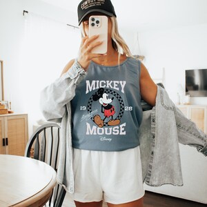 Pu&ograve; includere: Canotta blu con grafica di Topolino, la scritta "Mickey Mouse" e "1928". L'outfit comprende pantaloncini bianchi e una giacca di jeans. Viene indossato un berretto da baseball nero. L'immagine &egrave; stata scattata in interni.