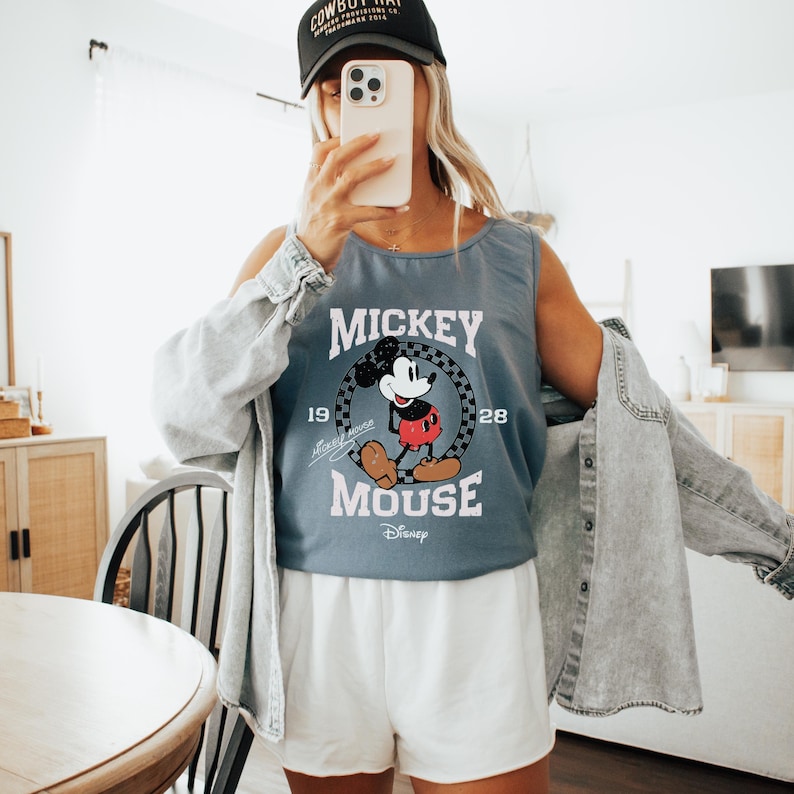 Pu&ograve; includere: Canotta blu con grafica di Topolino, la scritta "Mickey Mouse" e "1928". L'outfit comprende pantaloncini bianchi e una giacca di jeans. Viene indossato un berretto da baseball nero. L'immagine &egrave; stata scattata in interni.