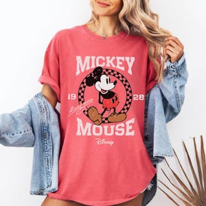 May include: A coral-colored t-shirt featuring a distressed graphic of Mickey Mouse. The design includes the words "Mickey Mouse" and "Disney", along with the year "1928". The shirt has short sleeves and a relaxed fit.