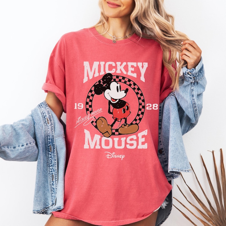 May include: A coral-colored t-shirt featuring a distressed graphic of Mickey Mouse. The design includes the words "Mickey Mouse" and "Disney", along with the year "1928". The shirt has short sleeves and a relaxed fit.