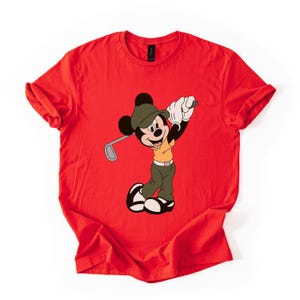 May include: Red t-shirt featuring a cartoon Mickey Mouse character in a golf outfit, swinging a golf club. Mickey is wearing a green hat, khaki pants, and a yellow shirt. The shirt is a solid red color.