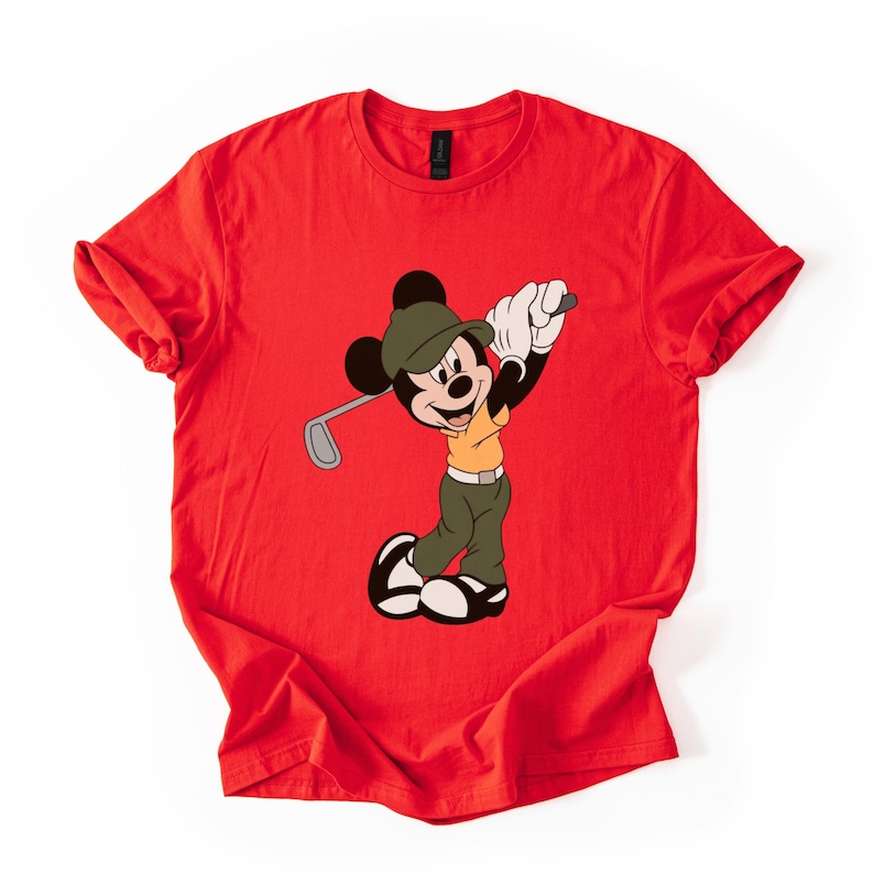 May include: Red t-shirt featuring a cartoon Mickey Mouse character in a golf outfit, swinging a golf club. Mickey is wearing a green hat, khaki pants, and a yellow shirt. The shirt is a solid red color.