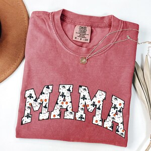 May include: A dusty rose Comfort Colors t-shirt with the word "MAMA" in a white, patterned font. The pattern includes bats, witch hats, and pumpkins. A silver necklace with a small pendant is draped across the shirt.