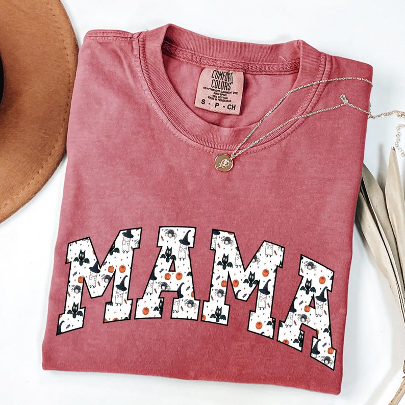 May include: A dusty rose Comfort Colors t-shirt with the word "MAMA" in a white, patterned font. The pattern includes bats, witch hats, and pumpkins. A silver necklace with a small pendant is draped across the shirt.