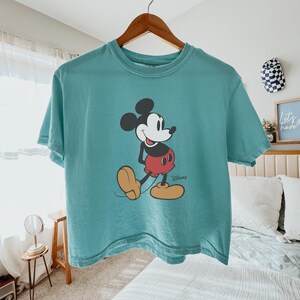 May include: Teal-colored t-shirt featuring a graphic of Mickey Mouse. The cartoon character is depicted in his classic pose, wearing red shorts, yellow shoes, and black gloves. The Disney logo is printed below the character.