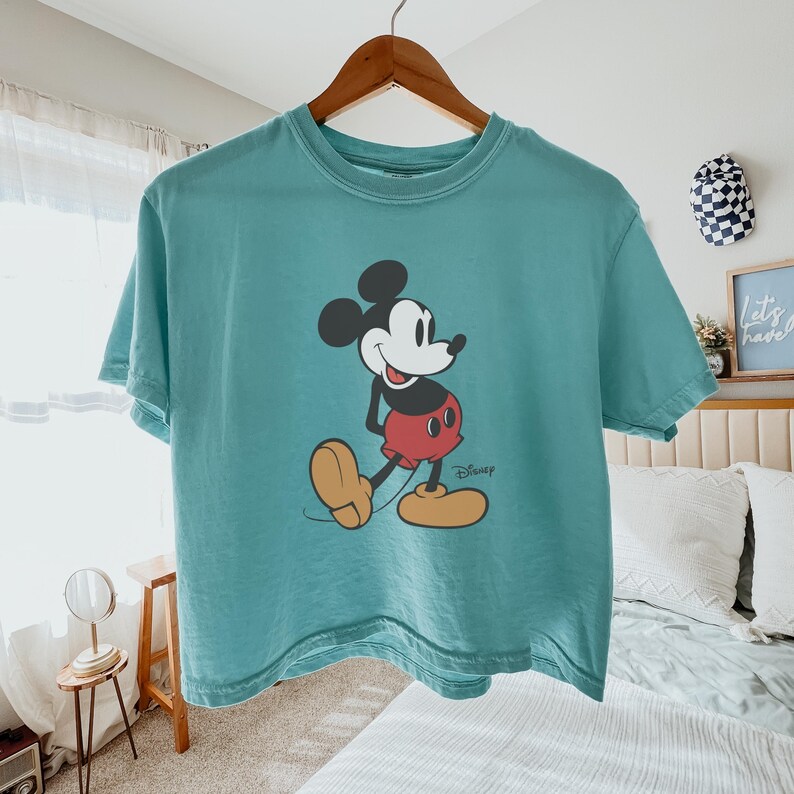 May include: Teal-colored t-shirt featuring a graphic of Mickey Mouse. The cartoon character is depicted in his classic pose, wearing red shorts, yellow shoes, and black gloves. The Disney logo is printed below the character.