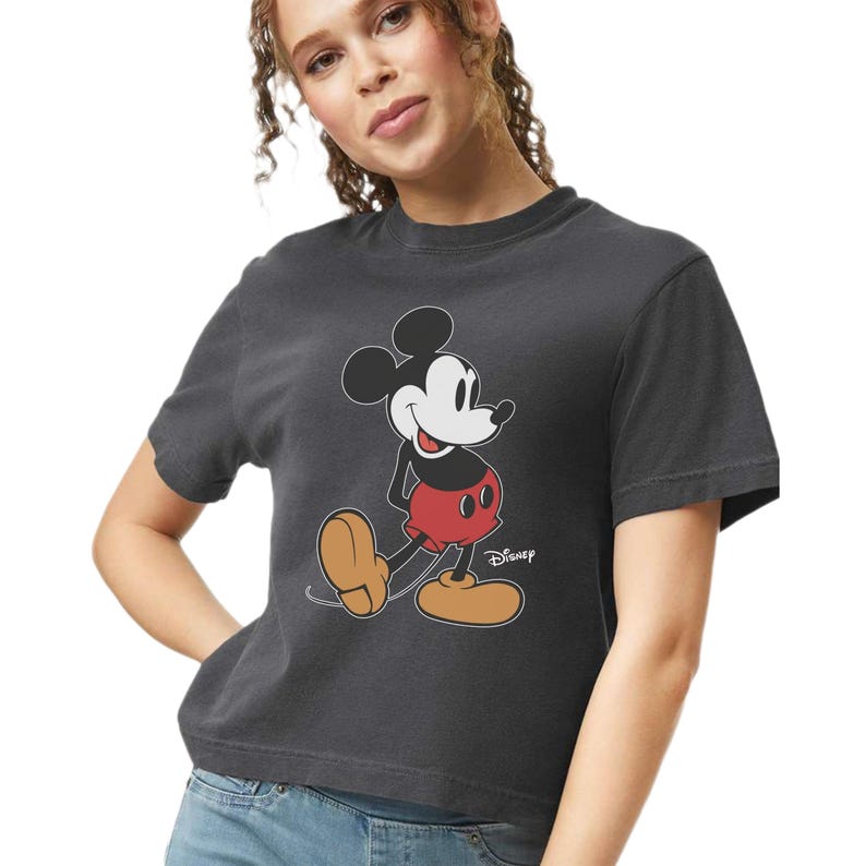 May include: Dark gray t-shirt featuring a classic Mickey Mouse graphic. The cartoon character is depicted in his iconic red shorts, white gloves, and large yellow shoes. The Disney logo is printed on the bottom right.