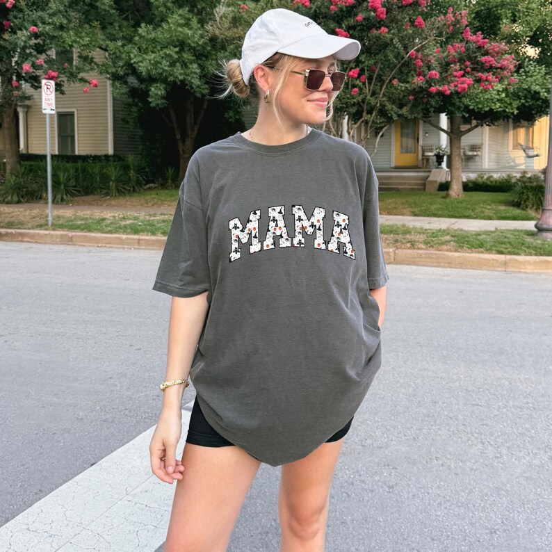 May include: Dark gray t-shirt with the word "MAMA" in white letters with a cow print design. The shirt is a relaxed fit and has short sleeves. The person is wearing a white baseball cap and sunglasses.