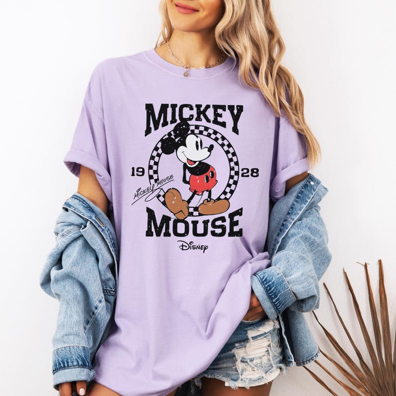 May include: A light purple t-shirt featuring a graphic of Mickey Mouse. The design includes the words "Mickey Mouse" and "Disney", along with the year "1928". The graphic is in black and white with red and brown accents.