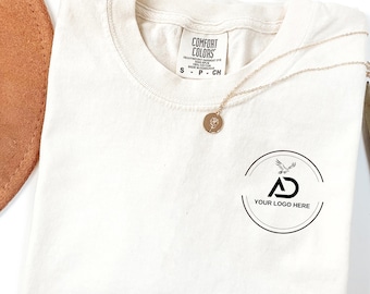 May include: Cream-colored Comfort Colors t-shirt with a round neckline, featuring a small logo on the front and a gold necklace. The shirt has a tag that reads "Comfort Colors". A brown hat is partially visible in the background.