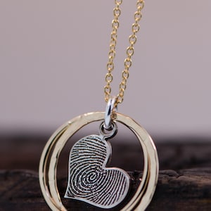 Puede incluir: Un collar de cadena dorada con un colgante de coraz&oacute;n plateado. El coraz&oacute;n tiene un dise&ntilde;o de huella digital en el interior. El colgante est&aacute; rodeado por dos anillos dorados.