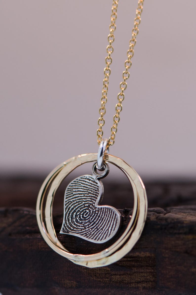 Puede incluir: Un collar de cadena dorada con un colgante de coraz&oacute;n plateado. El coraz&oacute;n tiene un dise&ntilde;o de huella digital en el interior. El colgante est&aacute; rodeado por dos anillos dorados.