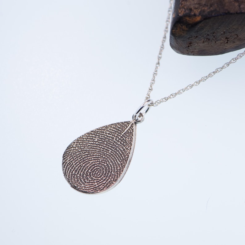 May include: A silver teardrop-shaped pendant necklace with a detailed fingerprint design. The necklace is on a silver chain.