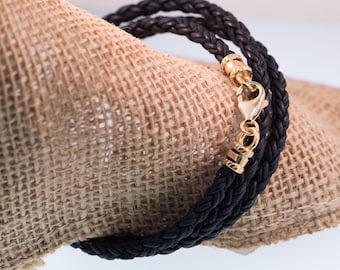 May include: A black braided leather bracelet with gold-colored accents and a clasp. The bracelet is wrapped around a tan burlap material, creating a rustic contrast. The bracelet appears to be made of three strands of braided leather.