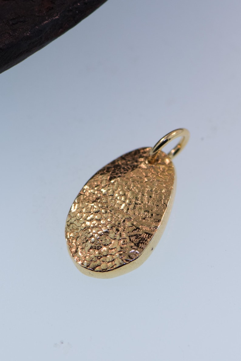 May include: A gold-toned oval pendant featuring a highly textured, cratered surface and a smooth, polished edge. The pendant includes a small, circular jump ring attached to the top, designed for threading onto a chain or cord for jewelry wear.