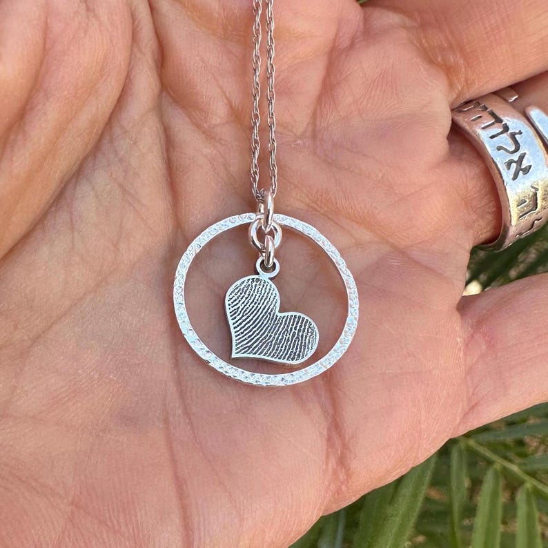 Puede incluir: Collar de plata con un colgante circular y un dije en forma de coraz&oacute;n. El dije en forma de coraz&oacute;n tiene un dise&ntilde;o de huella digital.