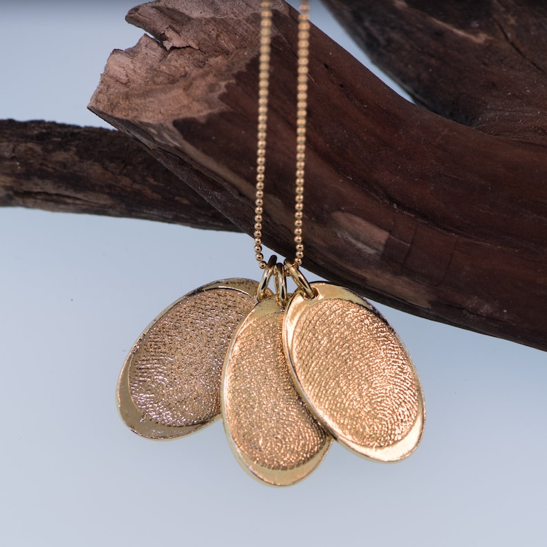 K&ouml;nnte beinhalten: Goldkette mit drei ovalen Anh&auml;ngern. Jeder Anh&auml;nger hat ein strukturiertes Design, das einer Fingerabdruck &auml;hnelt.