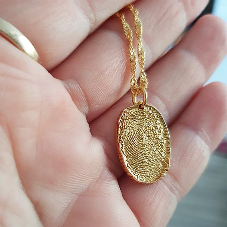 K&ouml;nnte beinhalten: Eine goldene Kettenkette mit einem Anh&auml;nger in Form eines Fingerabdrucks. Der Anh&auml;nger ist oval und hat eine detaillierte, strukturierte Oberfl&auml;che.