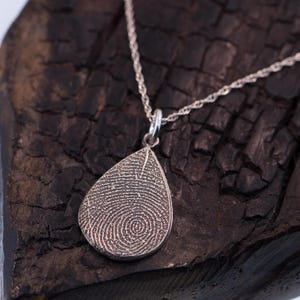 May include: A silver teardrop-shaped pendant necklace with a detailed fingerprint design. The necklace is on a dark wooden surface.