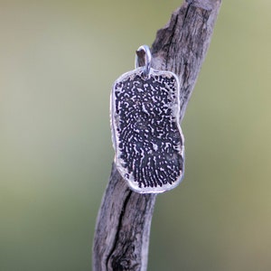 Peut inclure: Un pendentif en argent avec un motif d'empreinte digitale est suspendu &agrave; une boucle en argent. Le pendentif est rectangulaire avec des bords arrondis et une surface textur&eacute;e. Le pendentif est pr&eacute;sent&eacute; sur un fond vert flou.