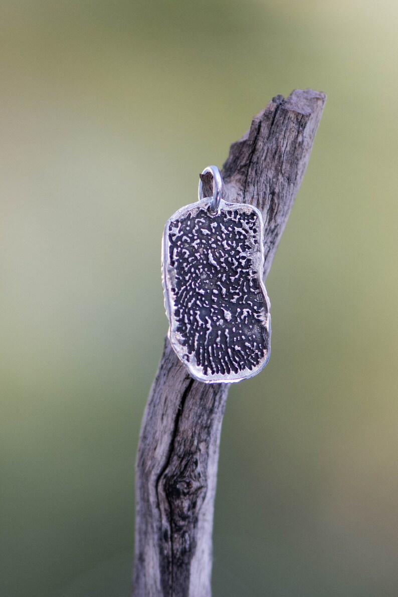 Peut inclure: Un pendentif en argent avec un motif d'empreinte digitale est suspendu &agrave; une boucle en argent. Le pendentif est rectangulaire avec des bords arrondis et une surface textur&eacute;e. Le pendentif est pr&eacute;sent&eacute; sur un fond vert flou.