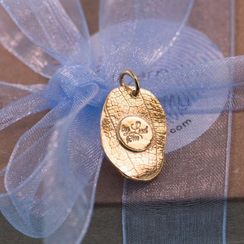 K&ouml;nnte beinhalten: Ein goldener Blattanh&auml;nger mit einer strukturierten Oberfl&auml;che und einem kleinen runden Anh&auml;nger mit der Inschrift "Joy Find Your Own" in einer kursivschriftlichen Schrift.